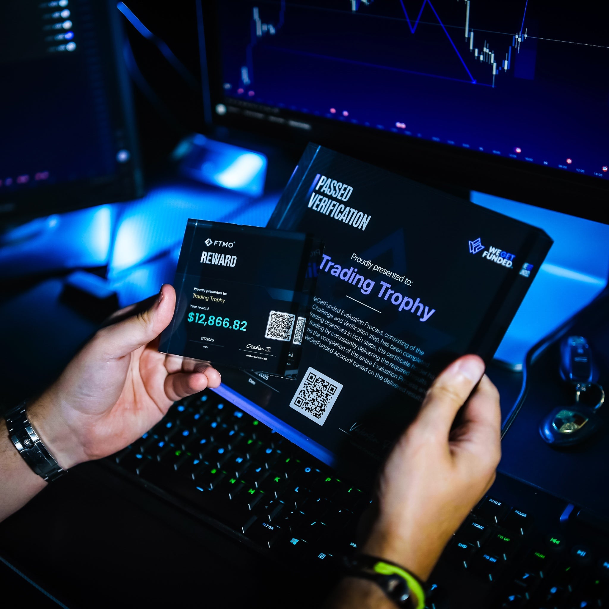 Two hands holding trophies in front of a computer setup with multiple monitors