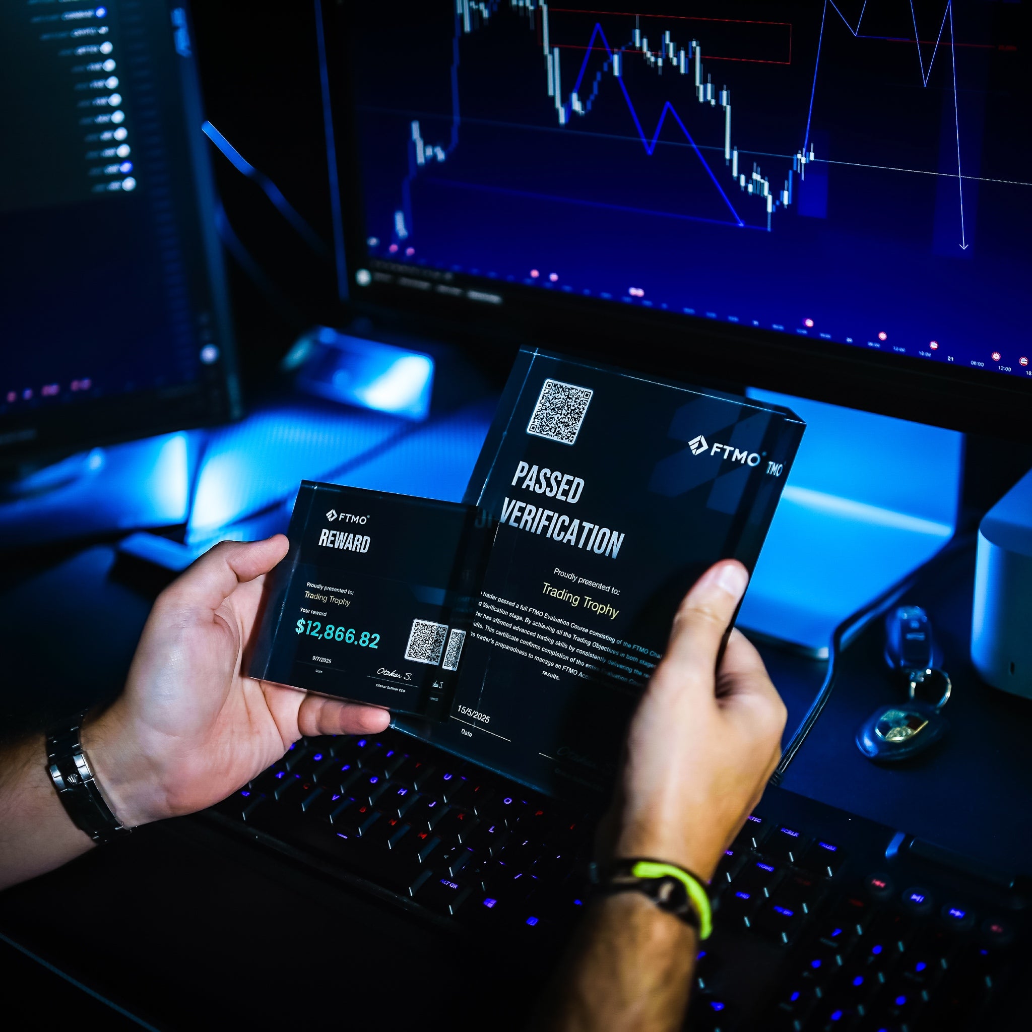 Two hands holding FTMO trophy in front of a computer screen with trading software